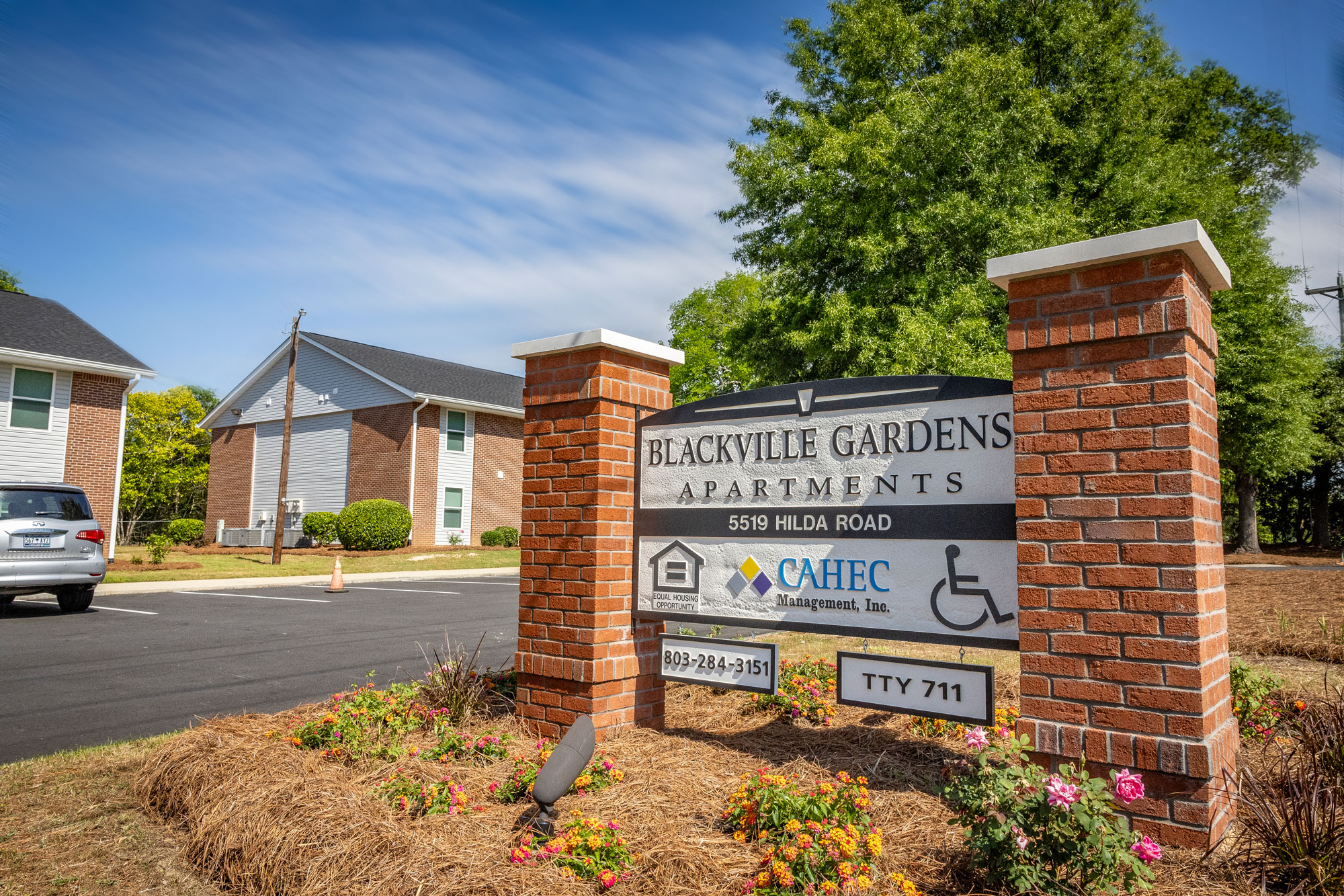 Sign in the foreground that reads "Blackville Gardens Apartments" with one of the apartment buildings in the background. Blue sky with few clouds in the background and a few green trees.