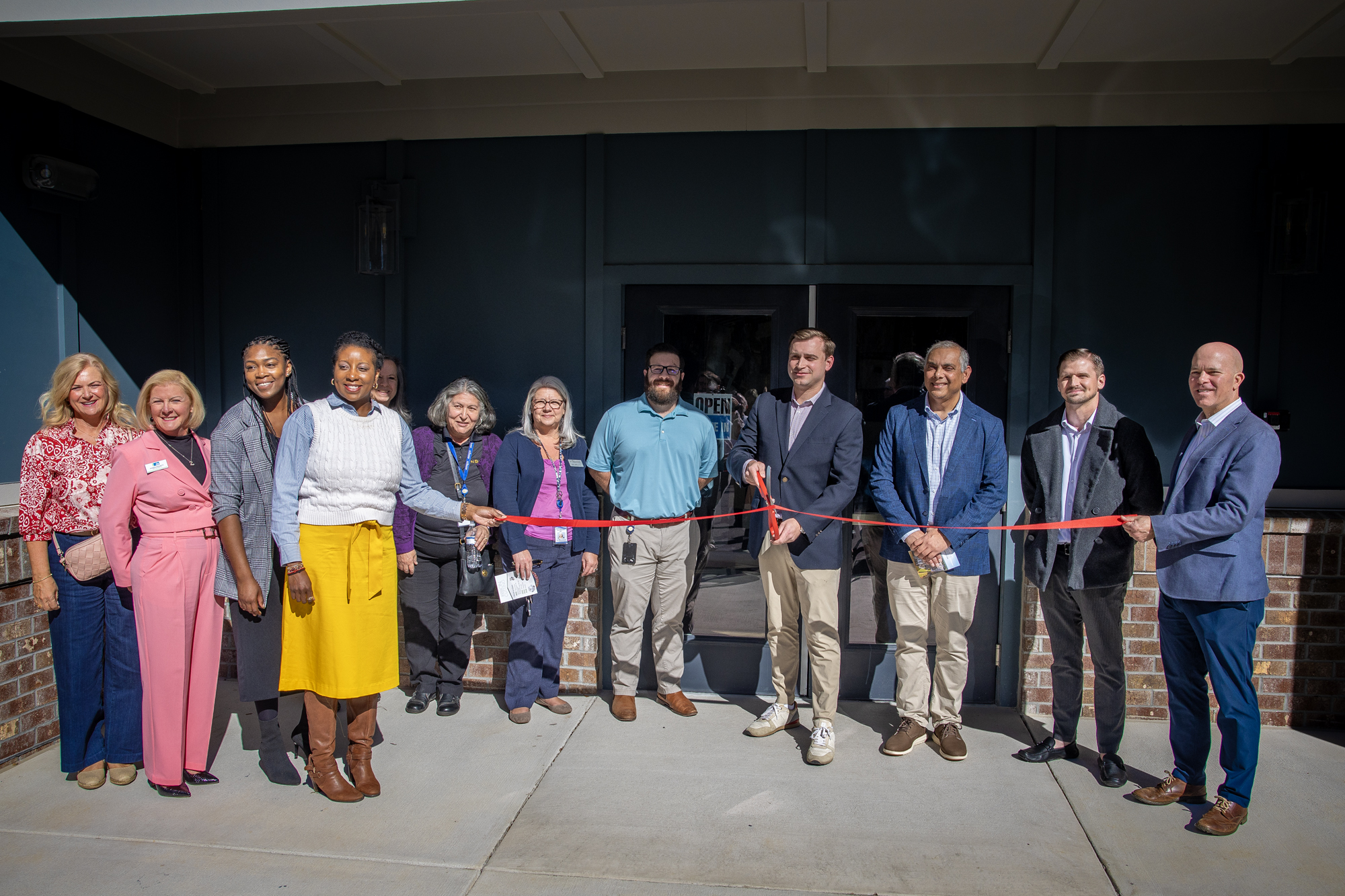 A group of more than a dozen people pose for a photo. All are in in business dress. Everyone is smiling. They're cutting a ribbon in front of an affordable housing development.